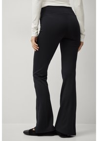 Schwarze, ausgestellte Hosen aus dehnbarem Stoff mit hoher Taille und glatter Textur. Die Details umfassen minimale Nähte und eine figurbetonte Silhouette.