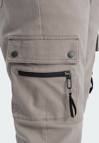 Beige cargobyxor med flera fickor, silverfärgade tryckknappar och en svart dragkedjeficka med en dragflik på lårområdet.