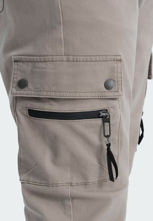 Beige cargo bukser med flere lommer, sølvfarvede trykknapper og en sort lynlåslomme med en trækfane på lårområdet.