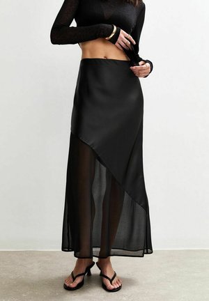 Jupe asymétrique noire en satin et tissu transparent, avec une texture lisse et une fente sur le côté. Associée à des sandales noires à lanières.