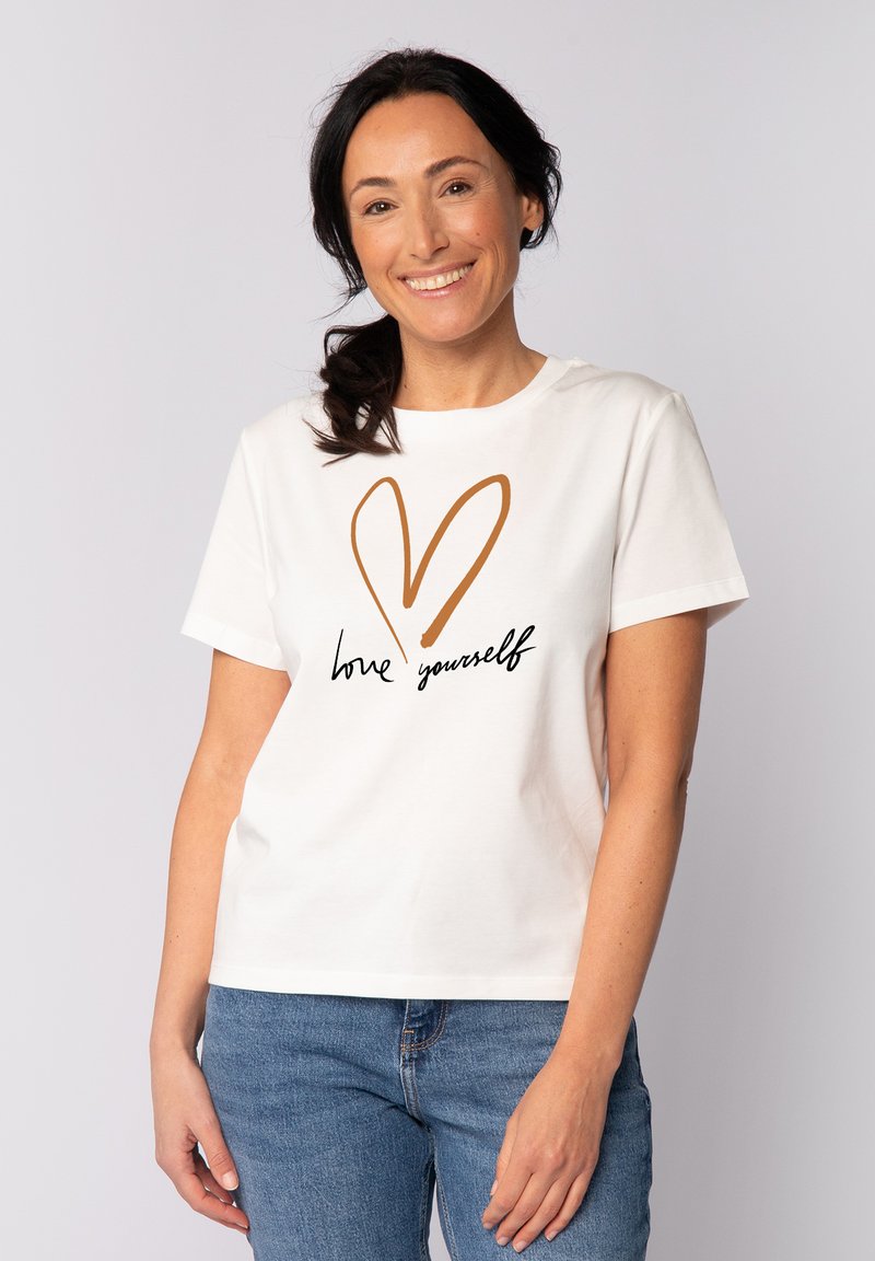 Witte katoenen t-shirt met korte mouwen, voorzien van een bruine hartomtrek en de tekst "hou van jezelf" in zwarte cursieve letters.