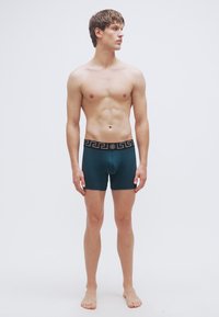 Βερμούδες μποξέρ σε τιρκουάζ με μαύρη μέση που διαθέτει γεωμετρικό σχέδιο. Το υλικό φαίνεται λείο και εφαρμοστό, κατάλληλο για καθημερινή χρήση.