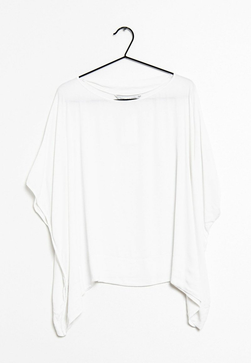 Blusa bianca sovradimensionata realizzata in un tessuto leggero. Presenta una scollatura ampia, spalle scese e un orlo asimmetrico. Appesa a un gancio nero.