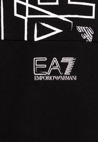 T-shirt nera in cotone con motivi geometrici e loghi bianchi. La scritta recita "EA7 EMPORIO ARMANI" in un font audace e moderno.