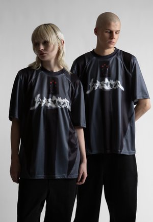 Két fiatal, akik sötét, túlméretezett ingeket viselnek, fehér graffiti feliratokkal és piros csillag részletekkel, világosszürke háttér előtt állnak.