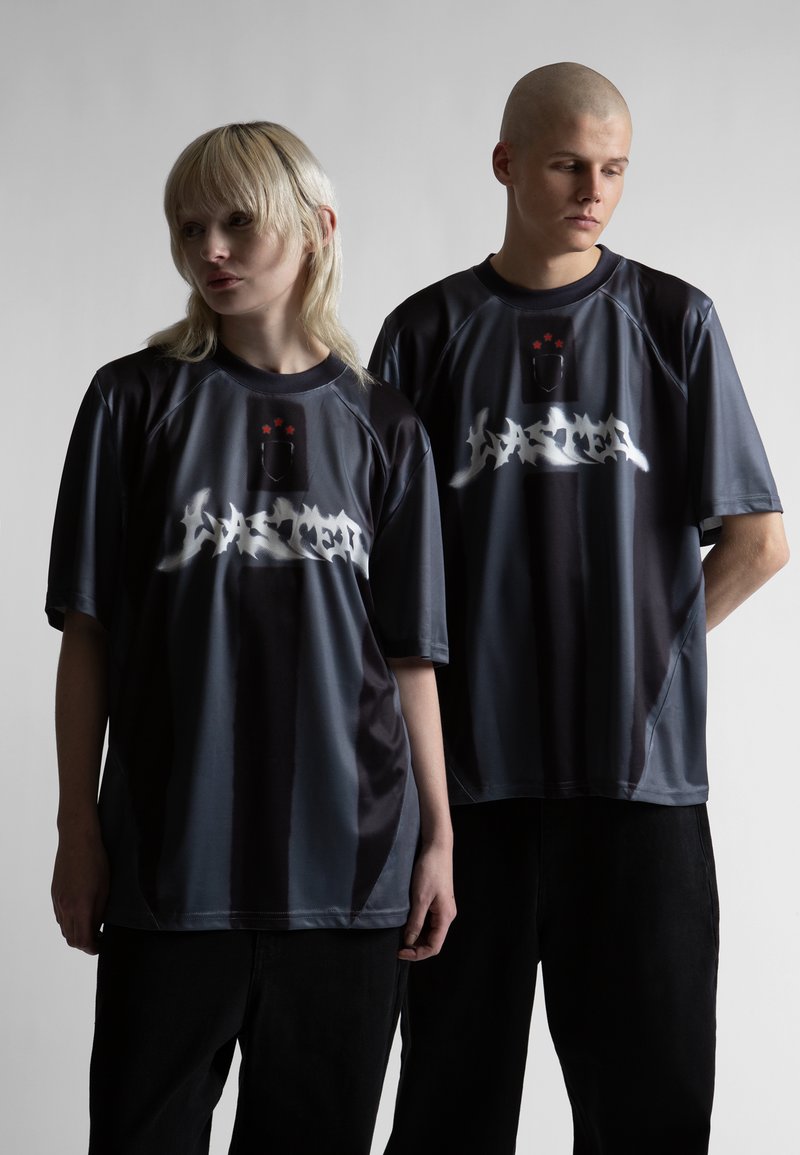 Δύο νέοι ενήλικες φοράνε ίδιου στυλ σκοτειρά φαρδιά μπλουζάκια με λευκό γκράφιτι κείμενο και κόκκινες λεπτομέρειες αστεριού, στέκονται μπροστά σε γκρι ανοιχτό φόντο.