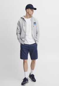 Hoodie zippé gris avec logo, t-shirt blanc, shorts cargo navy, chaussettes blanches et baskets noires. Casquette de baseball en navy. Tenue décontractée.