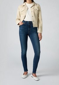 Beige džínsová bunda se dvěma náprsními kapsami, spárovaná s tmavě modrými skinny džíny. Model má na sobě bílé baleríny s černými akcenty.