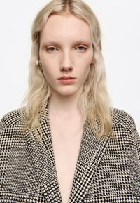 Persona joven con piel pálida y cabello rubio largo y ondulado, que lleva una chaqueta de pata de gallo en blanco y negro, mirando hacia adelante, sobre un fondo blanco.