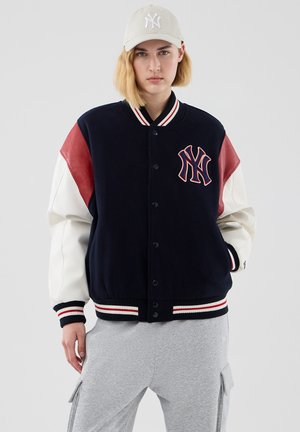 Person iført navy, hvid og rød varsity-jakke med NY-logo, grå cargobukser og en beige NY baseballkasket mod en ensfarvet baggrund.