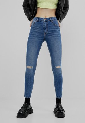 Femme portant un jean skinny bleu déchiré à taille moyenne, des chaussures noires épaisses, des chaussettes noires, un haut court vert citron et une veste noire sur fond gris.