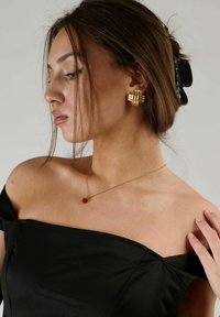 Guld geometriska örhängen och ett hjärtformat röd hängsmycke p�å en modell som bär en svart off-shoulder topp, slätt tygtextur.
