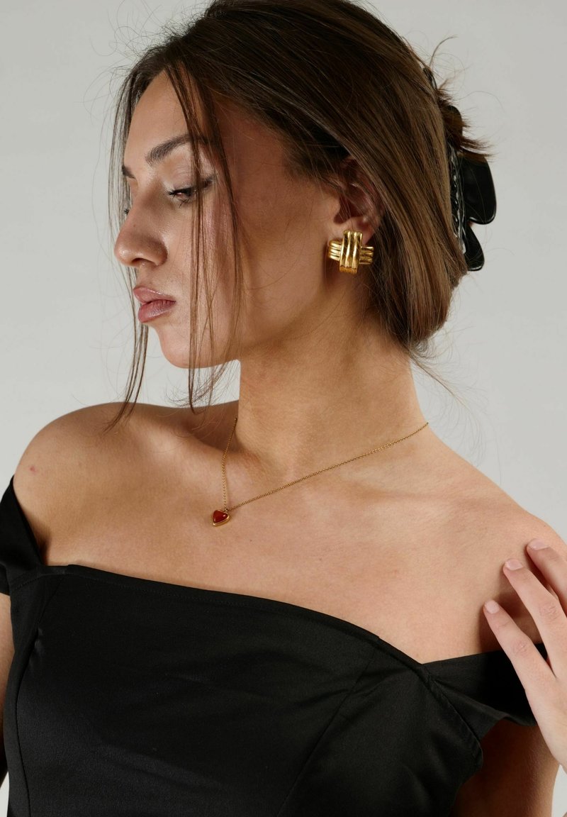 Guld geometriska örhängen och ett hjärtformat röd hängsmycke på en modell som bär en svart off-shoulder topp, slätt tygtextur.