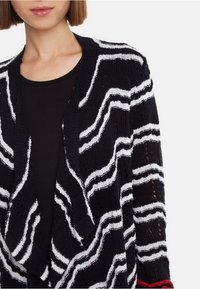 Cardigan lavorato a maglia a strisce bianche e nere con un motivo ondulato, apertura frontale e texture morbida; presenta un accento rosso sul polsino.