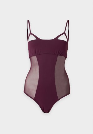 Femme debout portant un maillot de bain une pièce violet foncé à bretelles fines, assorti à des tongs noires, sur un fond clair uni.