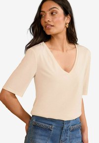 Beige top s výstřihem do V a krátkými rukávy, vyrobený z hladkého materiálu. Nosený s modrými denimovými šortkami s našitými kapsami a tkanými akcenty.