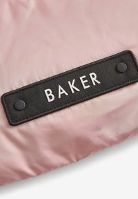 Rosa stofpose med et sort rektangulært mærke med teksten "BAKER" i hvid. Mærket har to runde sorte nitter til fastgørelse.