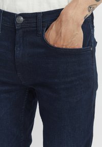 Jean en denim bleu foncé à coupe slim, avec un design classique à cinq poches, un dégradé subtil et un bouton en métal à la taille.