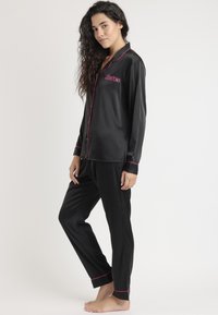 Conjunto de pijama de satén negro que presenta mangas largas, ribete rosa y bordado del logo "Barbie". Los pantalones tienen un diseño de pierna recta con dobladillos acampanados.