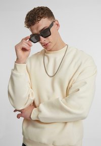 Kremasti oversized sweatshirt s teksturiranom tkaninom, rebrastim manžetama i srebrnim lancem. Crne naočale sa debelim okvirom.