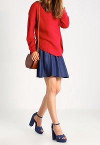 Rode gebreide trui, blauwe denim flared rok, blauwe hoge platform schoenen en een bruine schoudertas. Model staat tegen een neutrale achtergrond.