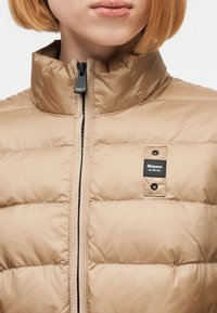 Κοντινό πλάνο ατόμου που φοράει μπεζ μπουφάν με επένδυση και ψηλό γιακά με φερμουάρ στο εμπρός μέρος, με ετικέτα "Blauer U.S.A." στο στήθος.