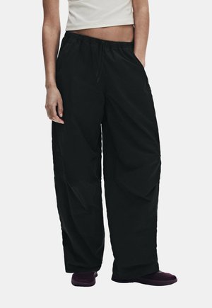 Personne portant un pantalon noir ample avec une ceinture élastique et un top blanc court, debout les mains dans les poches.