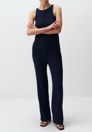 Woman wearing a sleeveless navy textured top and matching wide-leg pants with black open-toe heeled sandals, standing with arms crossed.