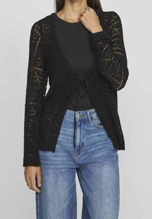Femme portant un cardigan noir crocheté sur un haut noir avec un jean bleu taille haute, la main près du cou, sur un fond gris uni.