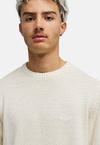 Nuori henkilö, jolla on lyhyet, vaalennetut hiukset, pukeutuneena kermanväriseen neulepuseroon, katsoo eteenpäin neutraali ilme kasvoillaan yksivärisen taustan edessä.