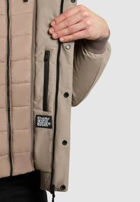 Világos bézs dzseki steppelt béléssel, cipzárral és patentos záródással, oldalzsebbel és textil címkével.