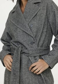 Schwarz-weißer Mantel im Fischgrätmuster mit doppelt geknöpften Knöpfen, einem breiten Kragen und einem gebundenen Taillengürtel. Texturiertes Wolle-Polyester-Gemisch.