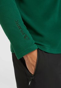 Grön fleece-tröja med en slät textur, prydd med en svart Polartec-logga på ärmen. Kombinerad med svarta byxor med dragkedja.