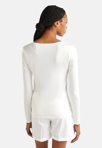 Vrouw met een gevlochten haar dat achteraan is vastgebonden, dragend een nauwsluitend wit lange mouwen shirt en witte korte broek met omslagen, van achteren bekeken.