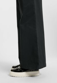 Black wide-leg pants paired with glossy black platform sneakers featuring white laces and a layered white sole with a serrated edge.