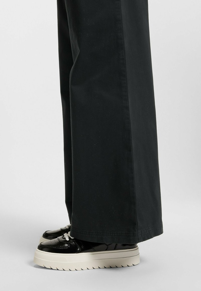 Black wide-leg pants paired with glossy black platform sneakers featuring white laces and a layered white sole with a serrated edge.