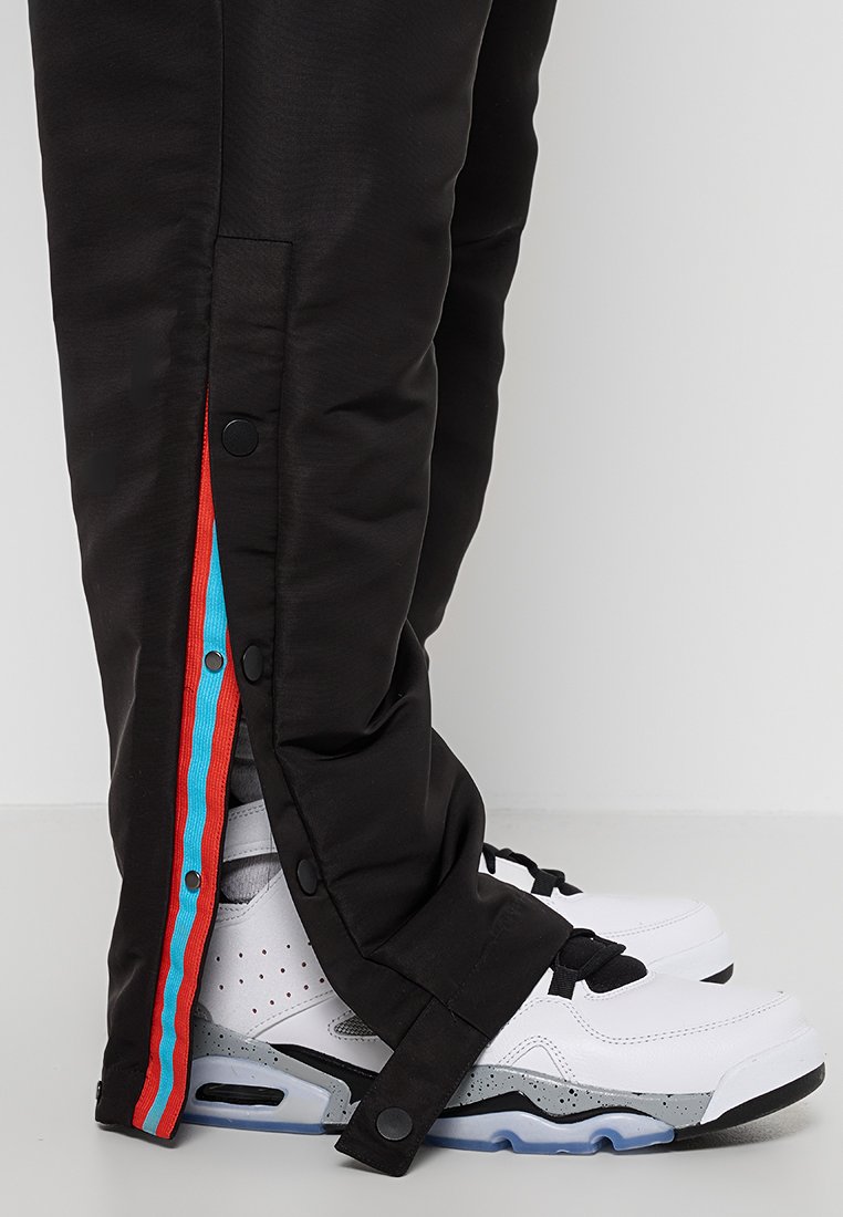 Schwarze Hosen mit Druckknopfverschlüssen und roten sowie blauen Akzenten an den Seiten. Kombiniert mit weißen Turnschuhen, die graue und schwarze Details aufweisen.