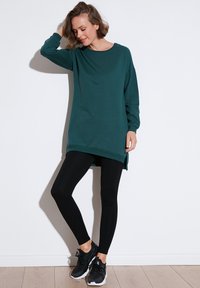 Türkiz oversized pulóver hosszú ujjakkal és oldalsó nyílásokkal, fekete leggingszel és fekete sportcipővel párosítva, fehér háttér előtt.