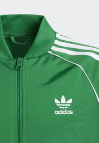 Zaļš adidas treniņjakas jaka ar stāvu apkakli, ar rāvējslēdzēju priekšā, trīs baltas svītras uz piedurknēm un baltu logo uz krūšu.