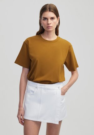 T-shirt en coton jaune moutarde à manches courtes et col rond. Porté avec une jupe en denim blanche avec une poche latérale et un bouton pression.