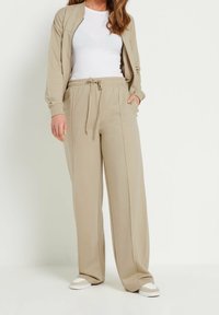 Conjunto de ropa de estar en beige que incluye una chaqueta con cremallera y pantalones holgados con cintura elástica. La tela es suave y tiene una textura lisa.