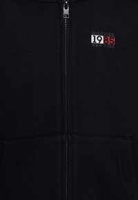 Schwarze Zip-Hoodie aus weichem Material mit einer vorderen Tasche, einem in Weiß und Rot gestickten Logo und kontrastierenden Nähten entlang des Reißverschlusses.