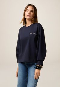 Giovane donna che indossa una maglietta a maniche lunghe blu navy con la scritta "Sport Chic" e jeans blu con decorazioni di perle sulle cosce.