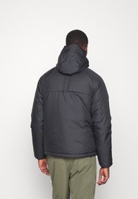 Svart vadderad jacka med huva, med en dragkedja på baksidan och en slät yta. Designen är enkel och funktionell.