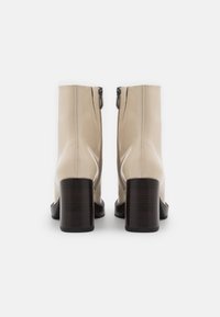 Botines de charol beige hasta el tobillo con puntera redonda y tacón cuadrado marrón. Cuentan con una cremallera lateral para poner y quitar fácilmente. Textura suave.