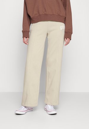 Persoon draagt beige wijde broek met "GAP"-logo op de heup, bruine cropped trui en witte lage sneakers op effen achtergrond.