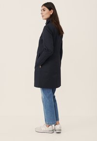 Marineblå lang frakke med høj krave, to sidelommer og lige snit. Båret over lyseblå jeans og grå sneakers. Glat tekstur.