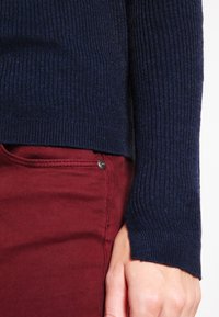 Granatowy sweter z prążkowanymi rękawami noszony z bordowymi spodniami, wyróżniający się gładką fakturą i dopasowanym krojem z delikatnym rozcięciem po bokach u dołu.