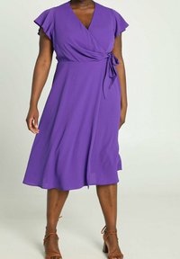 Femme portant une robe portefeuille midi violette avec des manches volantées et des sandales marron à talons blocs et à brides, debout devant un fond neutre.