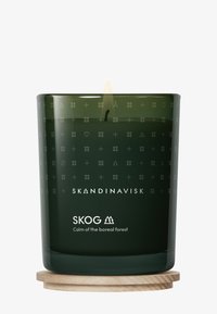 Groene glazen kaars met een gegraveerd patroon, brandende lont en houten basis. Etiket leest "SKOG Rust van het boreale woud."